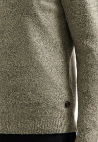 Close-up van een grijze gebreide trui met lange mouwen, gedragen over donkere pantalon, met een klein hexagonaal zwart label nabij de zoom.