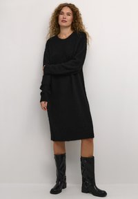 Juodas megztinis su ilgomis rankovėmis, apvalia kaklo linija ir raukšlėtomis rankovėmis. Suderinta su blizgiais juodais aukštos viršutinės dalies batais iki kelių.