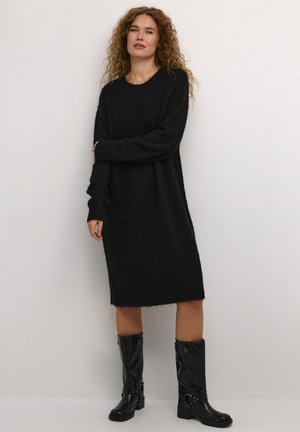 Robe pull en tricot noire avec des manches longues, un col rond et des poignets côtelés. Associée à des bottes noires brillantes montantes.