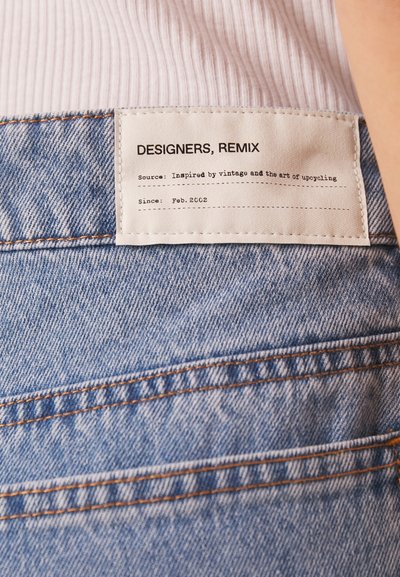 Dongeribukser med en falmet blå vask, med orange syninger. En hvid stoflabel viser mærket "DESIGNERS, REMIX" og plejedetaljer.