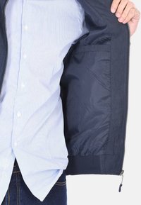 Giacca da marinaio con un'esterno liscio e un foderato interno lucido. Caratterizzata da una chiusura con zip e tasche aperte, indossata sopra una camicia a righe leggera.