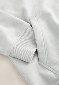 Hellgrauer Sweatstoff mit gerippten Bündchen; flache Nähte sichtbar, weiche Textur. Fokus auf Detail des Bündchens und glatte Oberfläche.