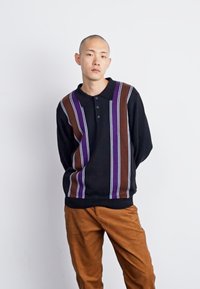 Pull noir avec des rayures verticales violettes et marron, doté d'un col et de trois boutons. Associé à un pantalon marron, texture lisse.