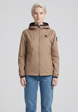 Giovane con capelli biondi corti che indossa una giacca beige con cappuccio e cerniera e jeans blu, in piedi contro uno sfondo neutro.