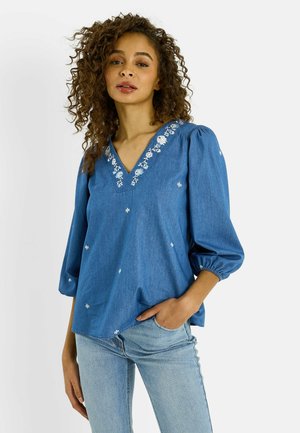 Mujer con cabello rizado que lleva una blusa azul con escote en V y bordado floral blanco, y unos vaqueros azul claro, posando con una mano en el bolsillo.