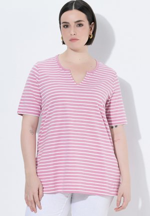 Donna con capelli corti e scuri indossa una maglia a righe rosa e bianca a maniche corte con scollo a V e pantaloni bianchi, in piedi contro un muro bianco.