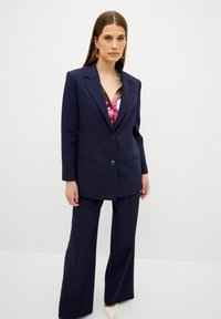 Námořnický oblek na míru s sakem na jeden knoflík, plochými kapsami a širokými kalhotami. Pod ním se nachází vícebarevná saténová košile s lesklým povrchem.