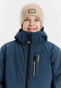 Blaue isolierte Jacke mit hohem Kragen und Reißverschlusstaschen. Beiger Strickmütze mit einem Logo-Patch, das Berge und Streifen zeigt.