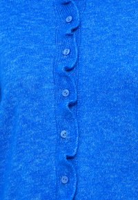 Cardigan bleu tricoté avec une texture douce, présentant un bord festonné le long de la patte de boutonnage et des boutons bleu clair.