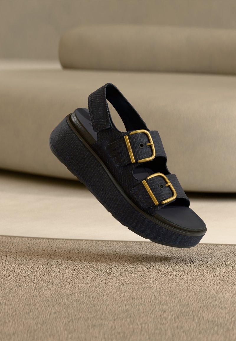 Μαύρο πλατφόρμα σανδάλι με δύο φαρδιές λουρίδες που διαθέτουν μεγάλες χρυσές αγκράφες, και ανοιχτή πλάτη, παρουσιάζεται να αιωρείται πάνω από μια μπεζ επιφάνεια.