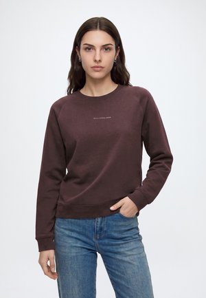 Donna con lunghi capelli castani che indossa una felpa marrone scuro e jeans blu, in piedi con una mano in tasca su uno sfondo semplice.
