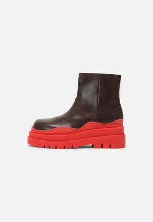 Bota de tobillo de cuero marrón oscuro con suela plataforma gruesa y ondulada de color rojo brillante y profunda banda de rodadura sobre un fondo gris claro.