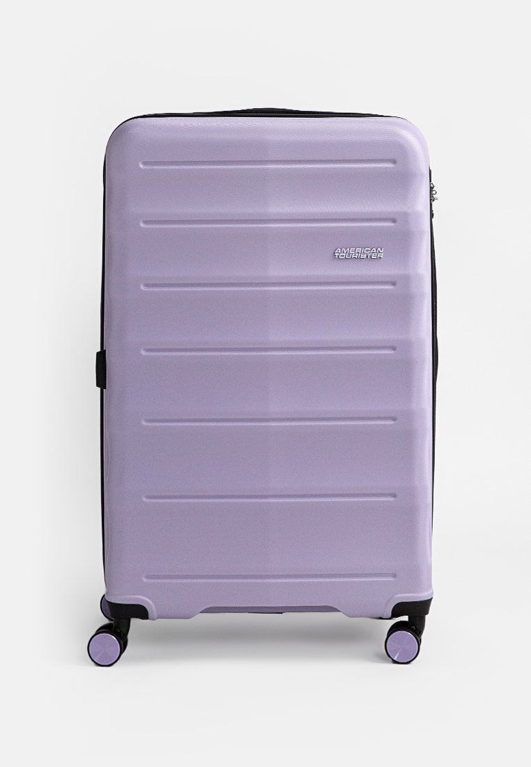 Valise rigide de couleur violet pastel, avec des rainures horizontales, une fermeture éclair, deux roues et un accent de fermeture éclair noir.