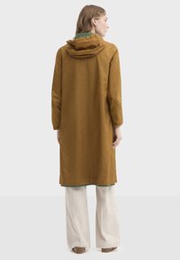 Longue veste marron imperméable avec capuche, en matériau léger, dotée de poignets élastiques et d'une doublure intérieure verte. Portée avec un pantalon large rayé.