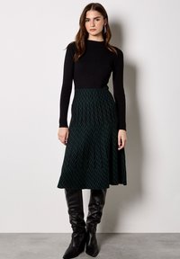 Donna che indossa una maglia nera a maniche lunghe a coste, gonna midi a fantasia verde scuro e stivali neri al ginocchio, in piedi contro un muro bianco.