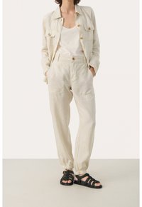 Jasno beżowa lniana kurtka z dwiema kieszeniami na piersi, w połączeniu z dopasowanymi spodniami z elastycznym mankietem. Uzupełniona czarnymi sandałami na paski.