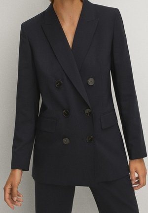 Persoon die een op maat gemaakte zwarte double-breasted blazer draagt met zes knopen en bijpassende zwarte broek tegen een neutrale achtergrond.