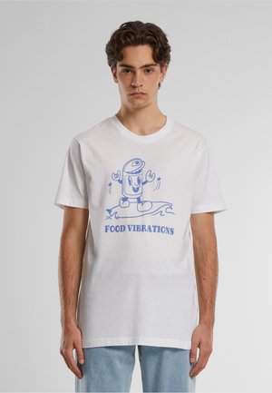 Młody mężczyzna w białej koszulce z niebieską kreskówkową puszką serfującą oraz napisem "FOOD VIBRATIONS" z przodu.