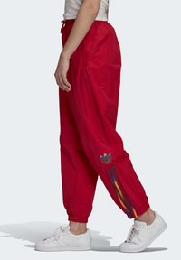 Červené sportovní kalhoty vyrobené z lehkého materiálu, s elastickým pasem, bočními kapsami a vícebarevnými pruhy po stranách.