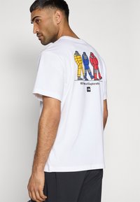 Λευκό βαμβακερό μπλουζάκι με πολύχρωμο γραφικό τύπωμα τριών φιγούρων με χειμερινά μπουφάν στην πλάτη, κείμενο από κάτω που γράφει «60 Χρόνια Εξερεύνησης».