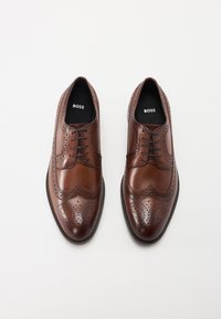 Brune lærdressko med dekorative brogue-detaljer, spiss tå og matchende snøring. Har en glatt tekstur og en kontrastfarge såle.