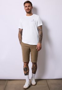 Bílý tričko s krátkým rukávem a modrou grafikou na levé části hrudníku, zkombinované s béžovými šortkami a bílými teniskami, doplněné o bílé kotníkové ponožky.