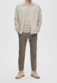 Veste chemise en lin beige clair, pull crème, pantalon à carreaux marron et chaussures claires. Tenue décontractée et texturée avec poches.