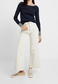 Marineblaues Langarmoberteil mit gestreiften Bündchen, kombiniert mit cremefarbenen, weit geschnittenen Hosen und beigen Wildledersneakern. Einfaches, lässiges Outfit-Design.