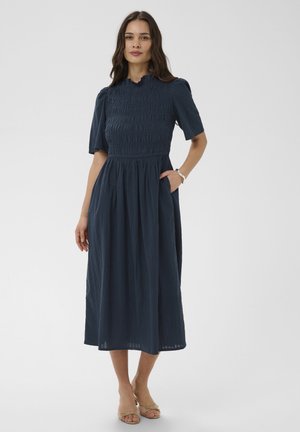 Femme debout, une main dans la poche, portant une robe midi bleu marine avec un corsage froncé et des manches courtes, des talons ouverts beiges.