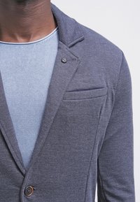 Mörkgrå kavaj med mjuk textur och en enkel knappstängning, med en snedskuren krage och bröstficka, lagd över en blå stickad tröja.