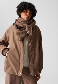 Giacca isolamento marrone con chiusura a scatto e polsini a coste, abbinata a una sciarpa spessa marrone. Texture del tessuto morbida, palette neutra.