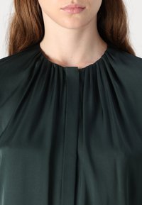 Mörkgrön, plisserad blus med hög krage. Slät, mjuk tyg med subtil lyster och en vertikal slits framtill för detalj.