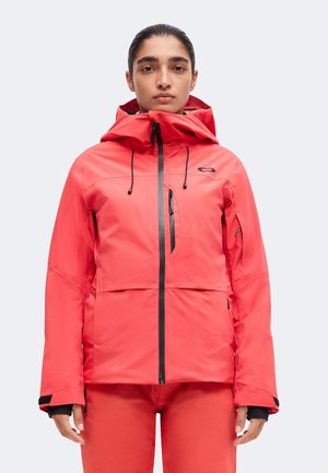 Personne portant une veste imperméable rouge vif avec une capuche, une fermeture éclair frontale et une poche poitrine zippée, debout devant un fond uni.