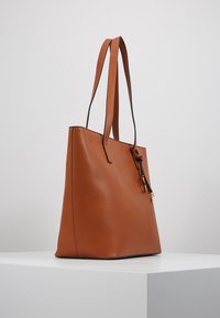 Hnědá kožená taška přes rameno s dvojitými popruhy a malým dekoračním přívěskem, umístěná na bílé ploše na jednobarevném pozadí.