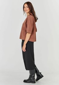 Tricou scurt, maro, cu mâneci largi, asortat cu pantaloni negri cu talie înaltă și cizme negre până la genunchi, pe un fundal neutr.