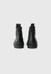 Bottes de cheville en cuir noir avec une texture lisse, inserts élastiques sur les côtés et une semelle en caoutchouc épaisse. La partie arrière présente des éléments de marque en doré.