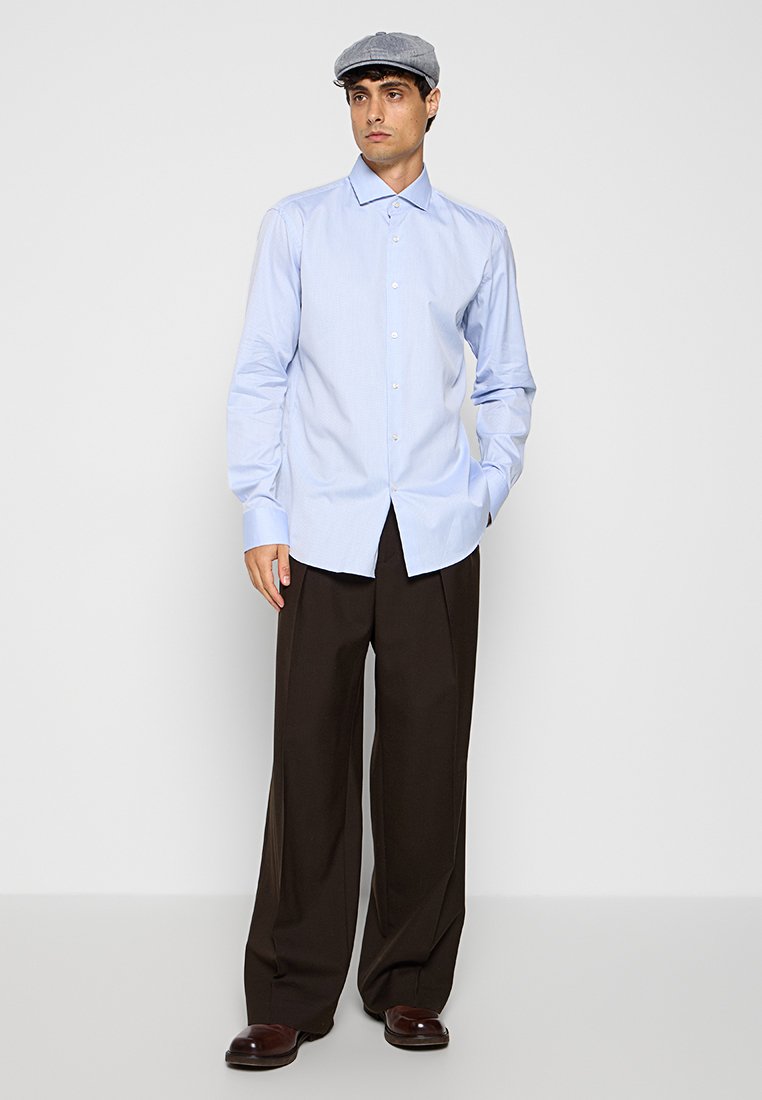 Lichtblauw overhemd met lange mouwen, gecombineerd met donkerbruine broek en een grijze platte pet, gedragen door een model tegen een effen achtergrond.