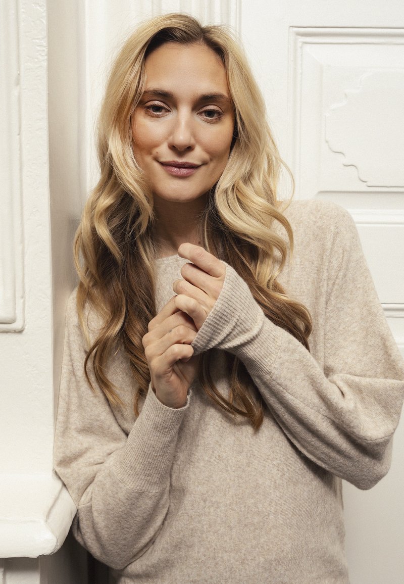 Beige stickad tröja med lös passform, ribbade muddar och mjuk textur. Modellen har vågigt blont hår och poserar mot en vit vägg.