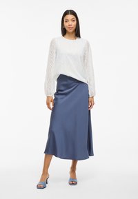 White textured blouse with long sheer sleeves, paired with a dark blue satin midi skirt. Model wearing light blue open-toe heels.