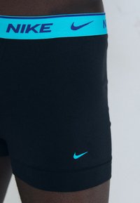 Schwarze Nike Boxer-Shorts mit einem blauen Bund, der den "NIKE"-Text und das Nike-Swoosh-Logo in Blau zeigt, aus glattem Stoff, in enger Passform.