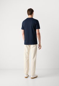 T-shirt blu navy a maniche corte con scollo rotondo, abbinato a pantaloni chiari e sneakers bianche, visto da dietro.