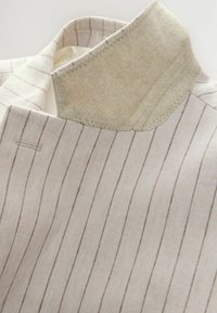 Helle beige maßgeschneiderte Jacke mit vertikalen braunen Streifen. Sie verfügt über einen weichen, beigen Filzkragen und saubere Nähdetails.