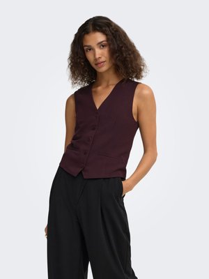 Donna con capelli ricci che indossa un gilet senza maniche bordeaux scuro abbottonato e pantaloni neri larghi, in piedi con una mano in tasca.