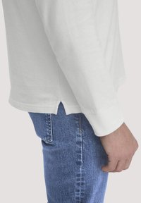 Hand rustend aan de zijkant, gekleed in blauwe jeans en een witte blouse met lange mouwen, voorzien van een manchet en een kleine zijspleet aan de onderkant.