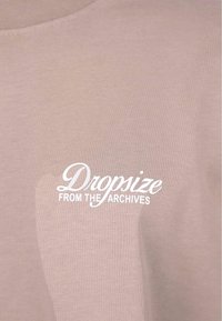 Hellbeige T-Shirt mit Rundhalsausschnitt, ausgestattet mit einem strukturierten Stoff und weißen Siebdrucktext: "Dropsize AUS DEN ARCHIVEN."