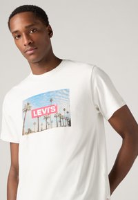 Tricou alb din bumbac cu un imprimeu colorat de palmieri și un cer albastru, având un logo roșu "LEVI'S" în centru.