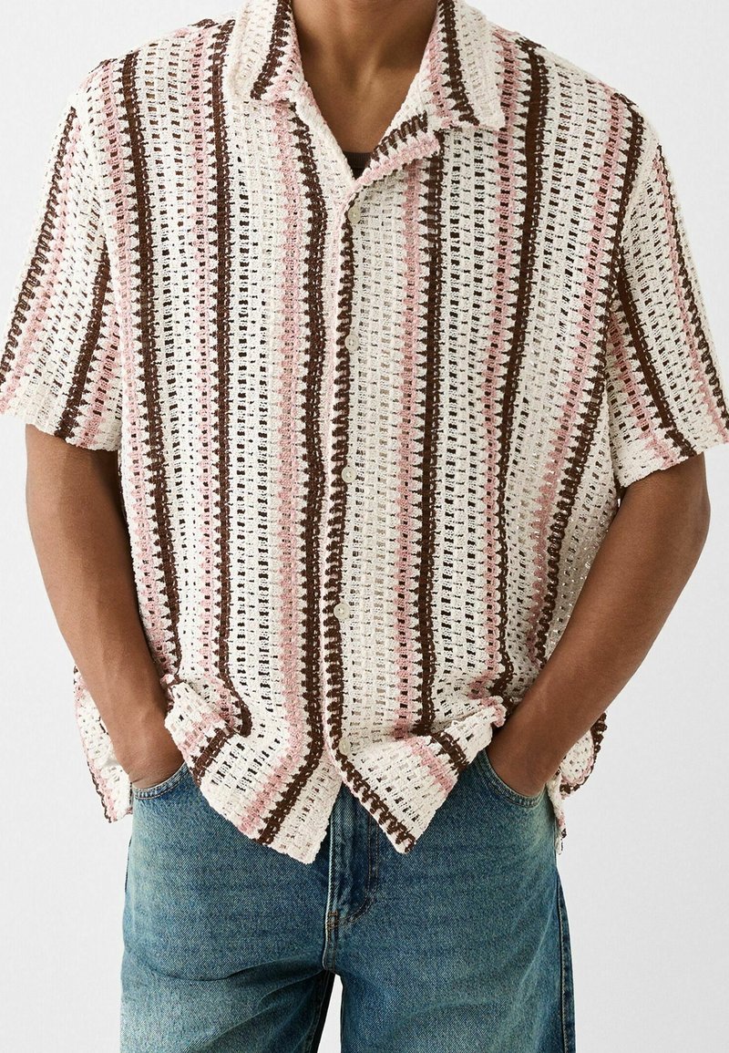 Homme portant une chemise en maille à manches courtes, boutonnée, avec des rayures verticales marron et rose, associée à un jean bleu, mains dans les poches.
