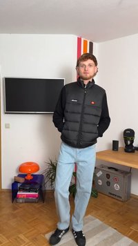 Melna pūkaina veste ar augstu apkakli, ar logotipu uz krūšu daļas, apvienota ar gaiši ziliem džinsiem un melniem sporta apaviem iekštelpu vidē.