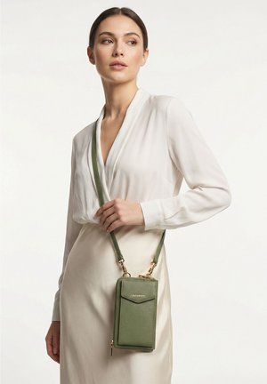 Mujer vestida con una blusa blanca y falda que lleva un bolso bandolera verde de cuero para teléfono con herrajes dorados y el logo "Lazarotti".
