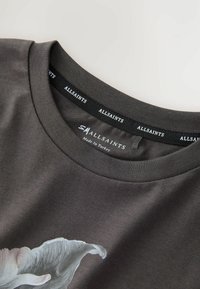 Detailný záber na tmavosivý výstrih trička AllSaints s potlačenou etikou a čiastočným bielym kvetinovým vzorom na látke.